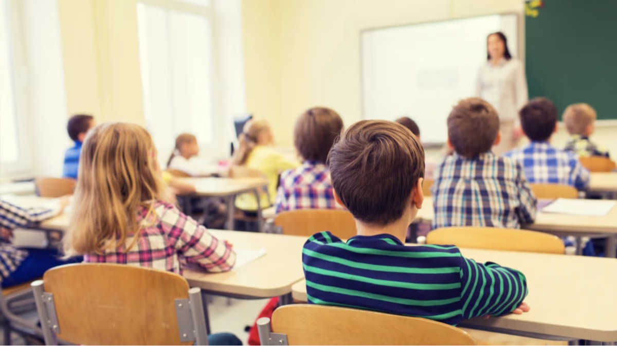 Salón de clases de primaria: alumnos en sus pupitres atendiendo a la maestra frente al pizarrón
